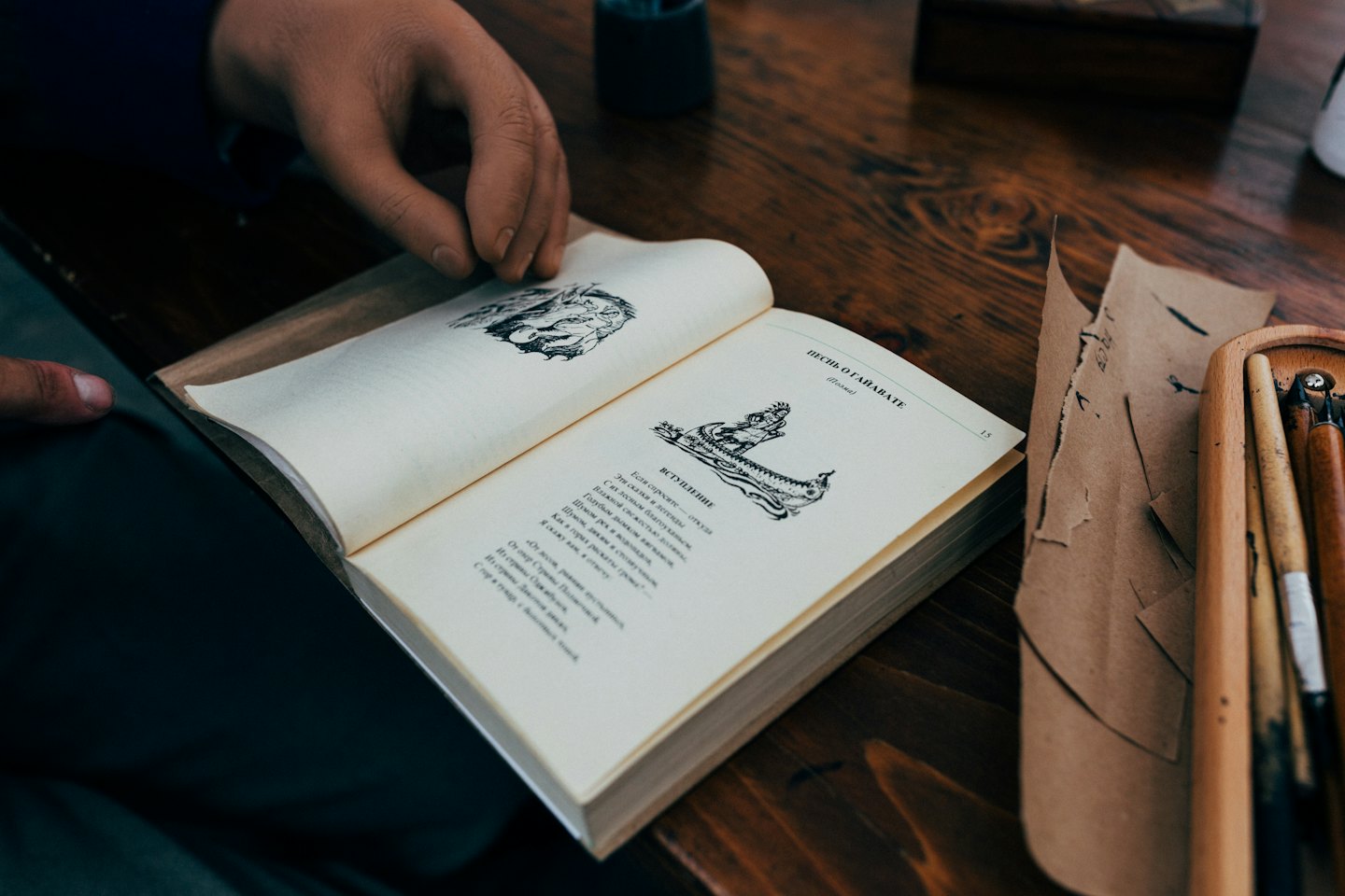 Hands holding open a book on a wooden table with art supplies.